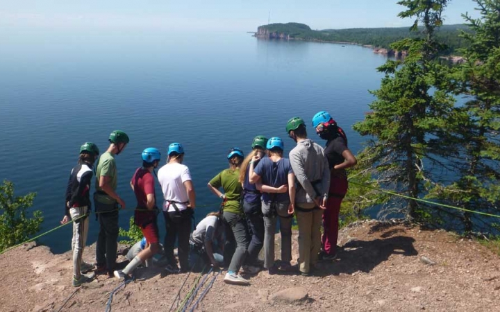 A group of students wearing safety gear stand on a rocky overlook above a vast blue lake. 